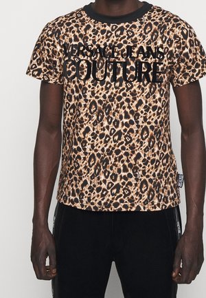 Mann trägt ein leopardgemustertes Versace Jeans Couture T-Shirt mit schwarzem Kragen und schwarze Hose mit seitlichen Logo-Streifen.