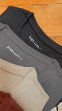 Three T-shirts stacked: black, gray, and cream. Each has a crew neck, short sleeves, and a smooth texture. The brand label is visible.