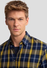 Patikrinta marškiniai mėlynos, geltonos ir pilkos spalvų tonais. Yra mygtukų apykaklė ir minkšta medžiaga, reguliaraus silueto. Išskirtinė plaid raštas.
