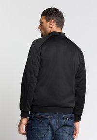 Zwarte bomberjack met een geribde kraag, manchetten en zoom. Gladde stof met een getailleerd ontwerp, voorzien van contrasterende stiksels en zonder zichtbare hardware.