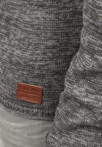 Maglione grigio a maglia con un motivo testurizzato e un'etichetta in pelle marrone con la scritta "BLEND" in rilievo. Mano parzialmente visibile, che mostra la manica.
