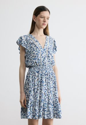 Egyenes barna hajú nő fehér ruhában, kék virágmintával, V-nyakkivágással, fodros ujjal és húzott derékkal, fehér háttér előtt áll.