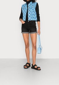 Blåmönstrad kort jeansväst, svart långärmad topp, högmidjade svarta shorts med uppvikt kant, svarta sandaler och en ljusblå texturerad handväska.