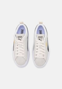 Puma-sneakers med en ljusbeige mockaöverdel, svarta accenter, vit sula och platta snörningar. Har varumärkeslogotyp på tungan och sidan.