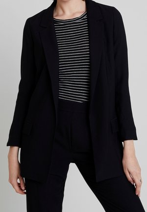 Persona che indossa un blazer nero sopra una camicia a righe bianche e nere con pantaloni neri su uno sfondo grigio semplice.