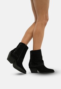 Botas de tobillo de ante negro con un tacón cónico, que presentan un detalle de tachuelas a lo largo del borde superior y una cremallera lateral para el cierre.