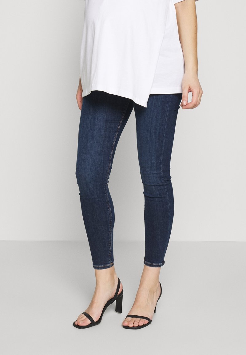 Jeans ajustados de color azul oscuro con una textura de denim suave y longitud hasta el tobillo, combinados con sandalias negras con tiras y tacones, y una blusa blanca suelta.