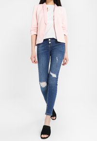 Světle růžový blazer přes bílý top, spárovaný s modrými úzkými džíny s odřenými detaily a černými skluzavkovými sandály.