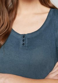 Šedý žebrovaný top s výstřihem do lodi, čtyřmi malými černými knoflíky na léze a jemnou, hladkou texturou.