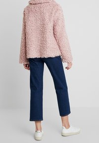 Růžová načechraná bunda s texturovanou tkaninou, volný střih, široké rukávy a límec, kombinovaná s tmavě modrými džíny a bílými teniskami.