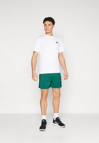 Weißes Sport-T-Shirt mit schwarzem Logo, kombiniert mit grünen Shorts. Das Modell trägt schwarze Sneakers mit weißen Akzenten. Schlichter Hintergrund.