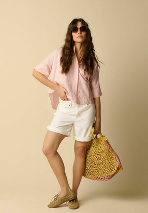 Frau trägt übergroßes hellrosa Hemd, weiße hochgekrempelte Shorts, Sonnenbrille, beige Loafer, hält eine gelb-rosa geflochtene Tasche vor beigem Hintergrund.