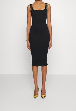 Kobieta ubrana w dopasowaną czarną sukienkę midi z ozdobnymi paskami i żółto-czarnymi szpiczastymi wysokimi obcasami, stojąca na tle jednolitego białego tła.