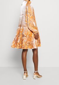 Orange swirling-patterned dress with a tiered hem, long sleeves, and gathered shoulders, made from lightweight fabric. Worn with beige block-heeled sandals.
