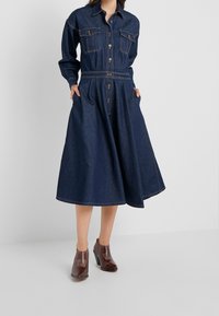 Dunkelblaues Jeanshemdkleid mit langen Ärmeln, durchgehender Knopfleiste, zwei Brusttaschen, tailliertem Schnitt und weit ausgestelltem Rock. Dazu gehören braune, hohe Stiefel.