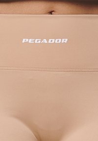 Närbild av en persons nedre torso i beige sömlösa underkläder med den vita "PEGADOR"-logotypen över midjebandet.