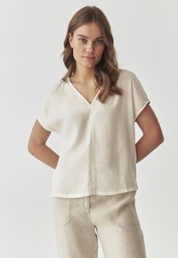 Blusa a maniche corte e scollo a V di colore crema chiaro, realizzata in un tessuto morbido e leggero. Presenta un orlo dritto e una vestibilità rilassata.
