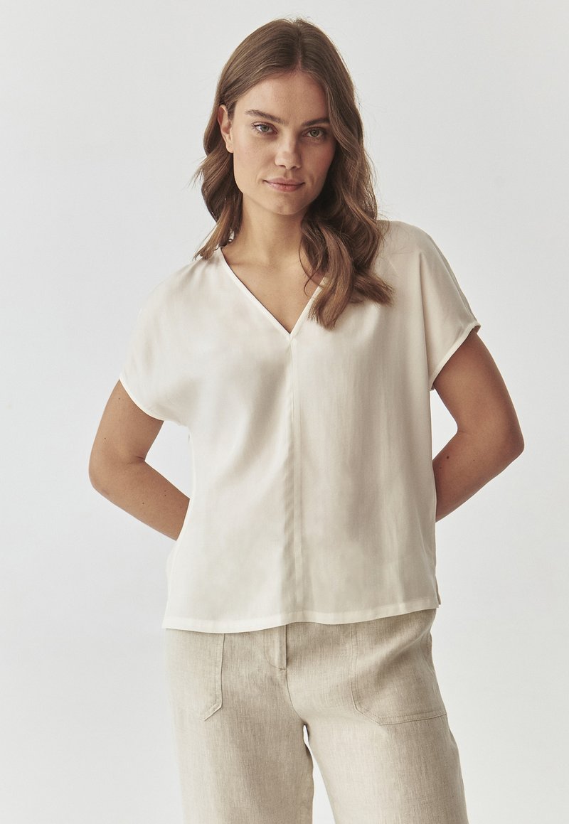 Blusa a maniche corte e scollo a V di colore crema chiaro, realizzata in un tessuto morbido e leggero. Presenta un orlo dritto e una vestibilità rilassata.