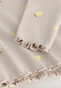 Jasnobeżowa żebrowana tkanina z żółtymi wzorami cytryn, ozdobiona falbankami na rękawach i dolnym brzegu, tworząc teksturę i detale.