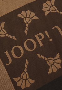 Pruun punutud tekstuur, millel on beežide lillemotiivide muster ja tekst "JOOP!" suurtes, julgetes kirjades. Sile tekstuur, ristkülikukujuline kuju.
