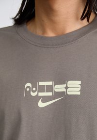 Nærbillede af en person iført en grå t-shirt med et stiliseret "Nike" logo i hvid, med en afslappet halsudskæring og en glat stoftekstur.