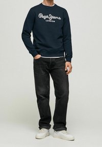Marinblå sweatshirt med grå "Pepe Jeans London" logotyp, rundad halsringning, ribbade ärmslut, matchad med mörka jeans och vita sneakers.