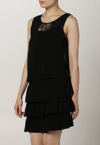 Musta kerroksellinen mekko, jossa on hihaton muotoilu, pitsikoristeet vartalolla ja rypytetty helma. Sujuva tekstuuri kauttaaltaan.