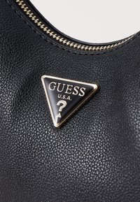 Materiale nero testurizzato con un emblema triangolare decorato in oro che riporta "GUESS U.S.A." e un punto interrogativo, vicino a una cerniera visibile.