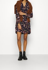 Květinové šaty v námořnické modři s dlouhými rukávy, výstřihem do V a volánkovým lemem. Hnědý svetr přehozený přes ramena. Černé kotníkové boty s silnými podrážkami.