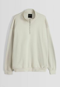 Pullover lungo a maniche lunghe color crema con colletto alto e zip a metà con un cursore metallico rotondo, polsini e bordo a costine.