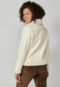 Tejfehér színű dzseki, texturált felülettel, műszőrme gallérral és mandzsettákkal. Barna kordbársony nadrág látható alatta. A modell hátsó nézete.