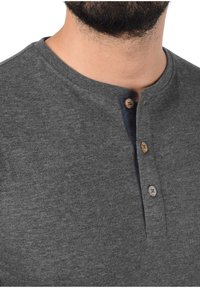 Man in een donkergrijze henley-shirt met drie gedeeltelijk ontbuttonde houten knopen en zichtbare halslijn en baardstoppels.