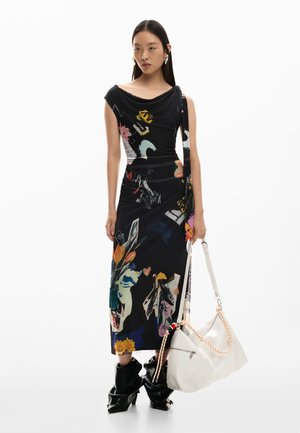 Vrouw in een zwarte midi-jurk met bloemenprint, zwarte slouchy laarzen, grote witte tas met oranje touwhandvatten, staand tegen een witte achtergrond.