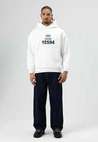 Witte hoodie met een zwarte opdruk in het midden met de tekst "POLIZEI 15594", gecombineerd met donkerblauwe wijde jeans en zwarte schoenen.