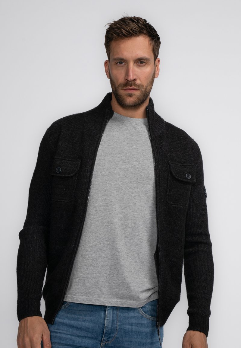 Zwarte ritssluiting cardigan van een textuur gebreide stoffen, met twee voorzakken met knopen, gedragen boven een lichtgrijs t-shirt en blauwe jeans.