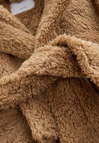 Weiches, sandfarbenes, dickes Fleece-Material mit dichter, flauschiger Textur und Falten, die Schatten und Tiefe erzeugen.