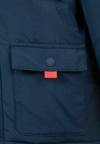 Navy blue jacket fabric with a textured finish, featuring a front pocket with a black snap button and a small red accent tab.