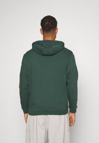 Hombre de espaldas, vistiendo una sudadera verde oscuro con la capucha bajada y pantalones cargo beige claro, frente a un fondo blanco liso.