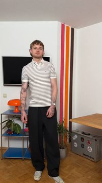 Νέος άνδρας με τατουάζ στέκεται σε ένα δωμάτιο με ριγέ τοίχους, τηλεόραση τοποθετημένη στον τοίχο, πορτοκαλί λάμπα, φυτά και μεταλλική θήκη αποθήκευσης.