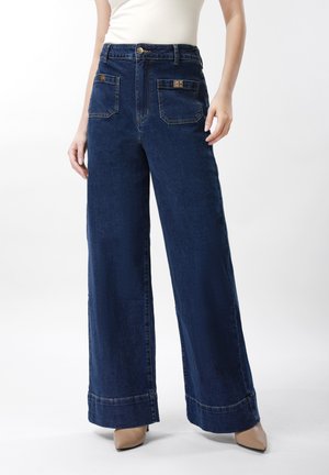 Nainen pukeutuneena korkeavyötäröisiin tummansinisiin leveälahkeisiin farkkuihin, joissa on etutaskut, ja beigen värisiin, kärjestä teräviin kenkiin, seisomassa valkoista taustaa vasten.