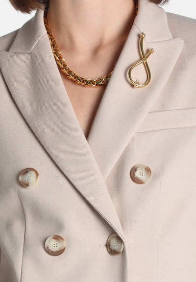 Femme portant un blazer beige croisé avec des boutons marron, un collier chaîne doré épais et une broche dorée de forme abstraite sur le revers.