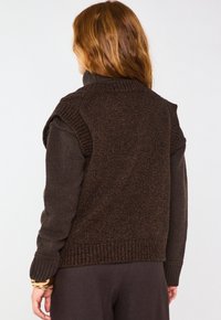 Gilet di maglia marrone con una finitura testurizzata, caratterizzato da un colletto alto e maniche corte, abbinato a una camicia a maniche lunghe scura sotto.