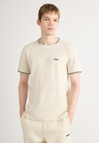 T-shirt en coton beige avec un col ras du cou, finitions noires au col et aux manches, arborant un petit logo "BOSS" noir sur la poitrine.