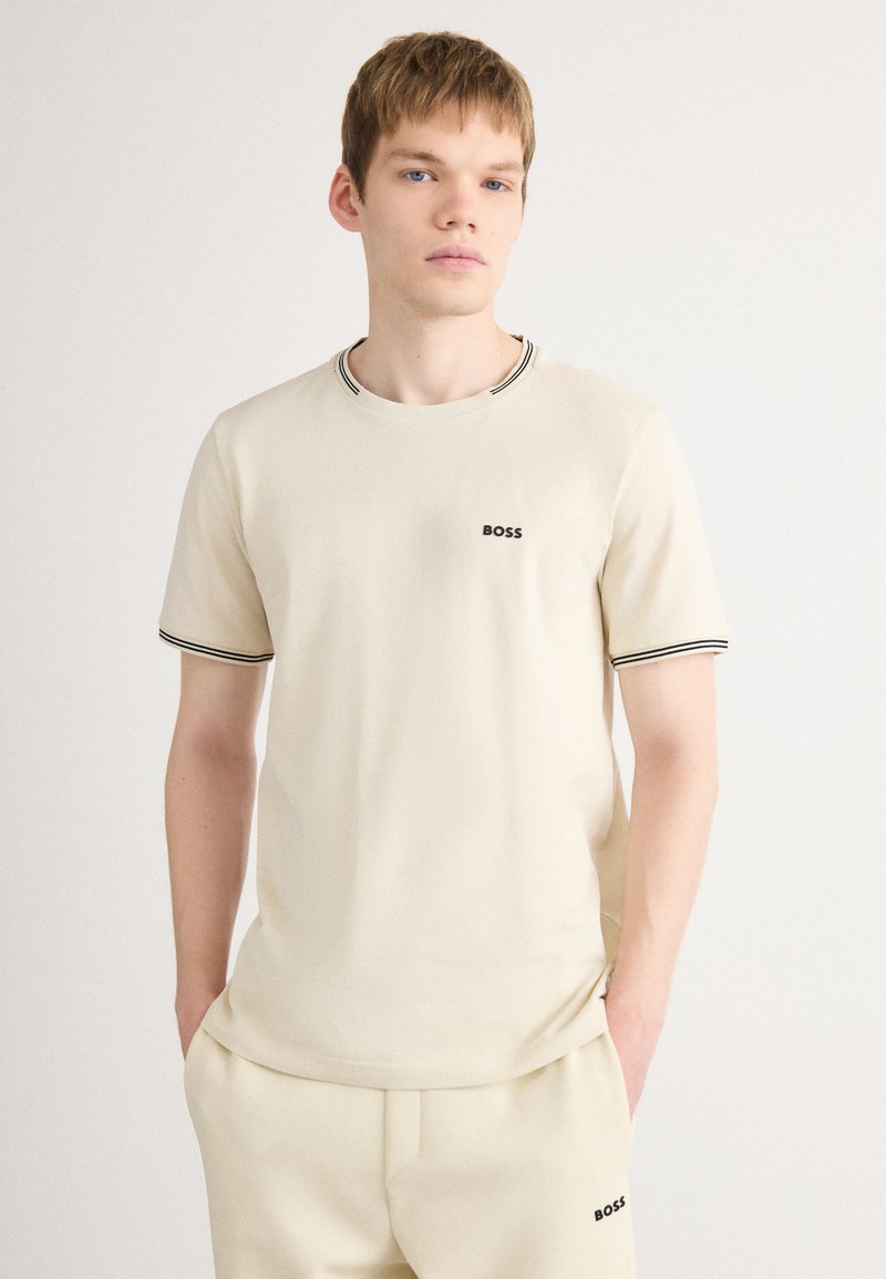 T-shirt en coton beige avec un col ras du cou, finitions noires au col et aux manches, arborant un petit logo "BOSS" noir sur la poitrine.