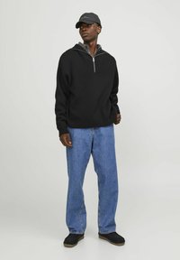 Schwarzer Fleece-Pullover mit Reißverschlusskragen, kombiniert mit ausgewaschenen blauen geraden Jeans und schwarzen Slippern, abgerundet mit einer dunklen Mütze.