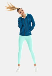 Noor naine türkiissinistes retuusides ja sinise lukuga kapuutsiga seisab, juuksed õhus lehvimas ja käed taskus, naeratades valge tausta ees.