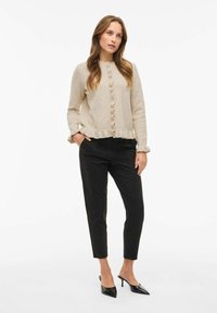 Bež pletena jopica z nabrano robom in gumbnim zapiranjem, v kombinaciji s črnimi ožjimi hlačami in črnimi salonarji s ostro konico.