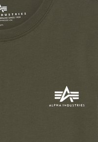 Camiseta verde oliva de algodón con cuello redondo. Presenta un logo blanco y el texto "ALPHA INDUSTRIES" cerca del pecho.