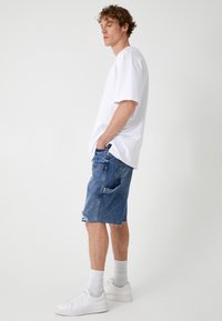 Camiseta blanca de manga corta, pantalones cortos de mezclilla azules con dobladillo deshilachado, combinados con zapatillas blancas y calcetas de longitud media, de pie contra un fondo claro.