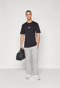 Černé atletické tričko s bílými pruhy na bocích, šedé tepláky s nenápadným logem a černá sportovní taška. Nošeno s bílými teniskami.